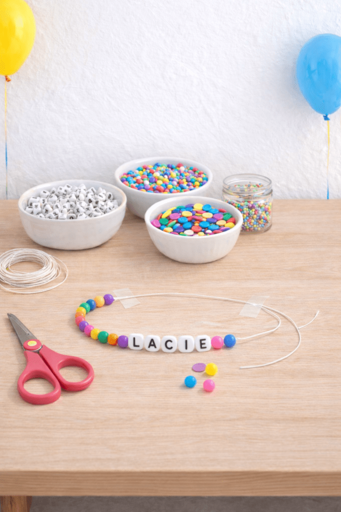Colorful DIY name bracelet with letter beads spelling Lacie on a wooden table with bowls of beads and string