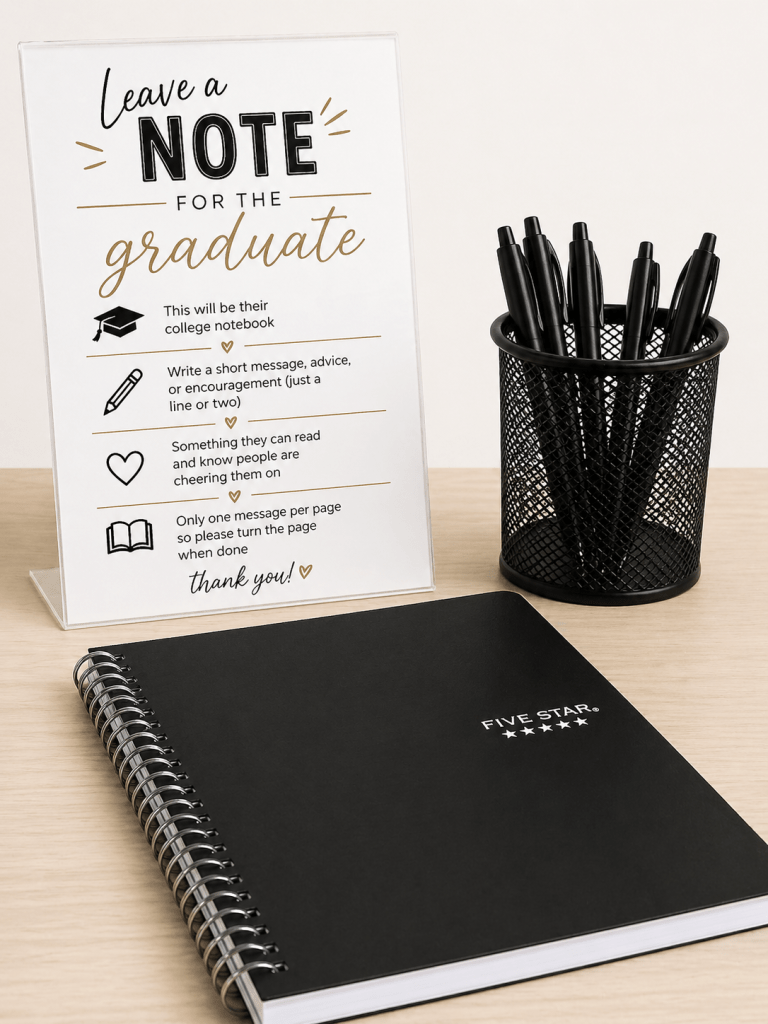 “Simple graduation party setup with a black Five Star notebook, black pens in a holder, and a small black-and-gold instruction sign inviting guests to leave notes for the graduate.”