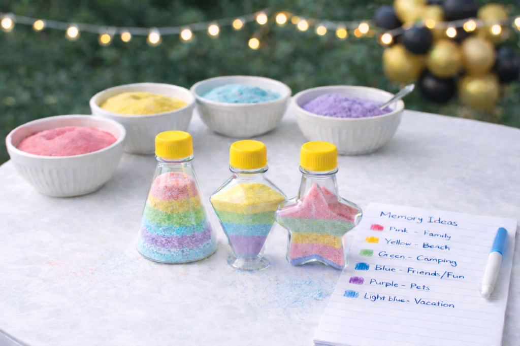 Graduation sand art memory jar activity setup on a white table with colorful sand, small glass bottles, and a graduate creating a layered sand jar outdoors