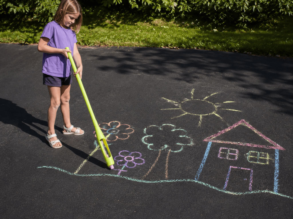 Young girl using a green stand-up sidewalk chalk holder to draw colorful flowers, trees, and a house on a blacktop driveway during outdoor summer play.